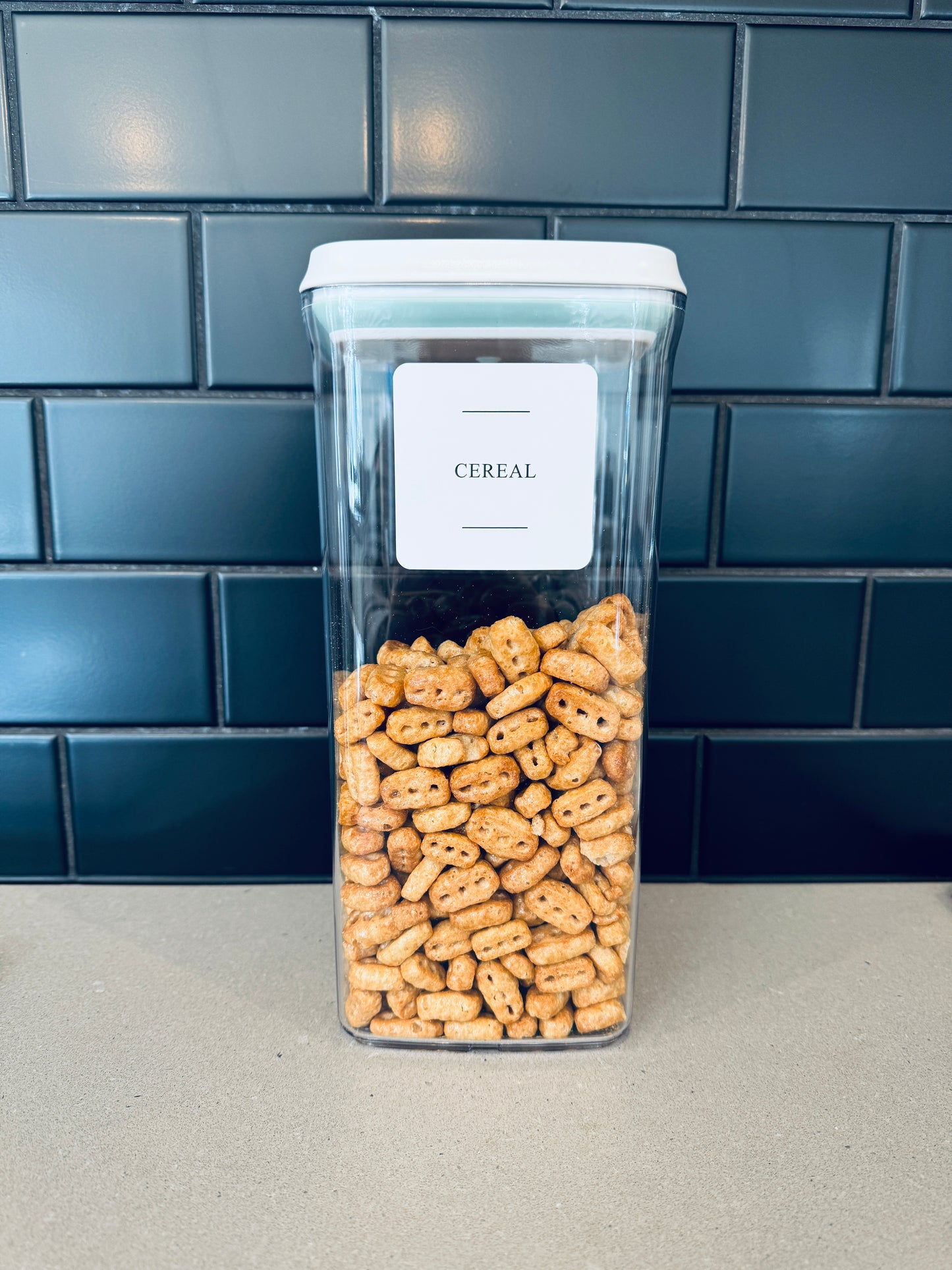 Clear container with cereal labeled 'Cereal' against a blue tiled wall.