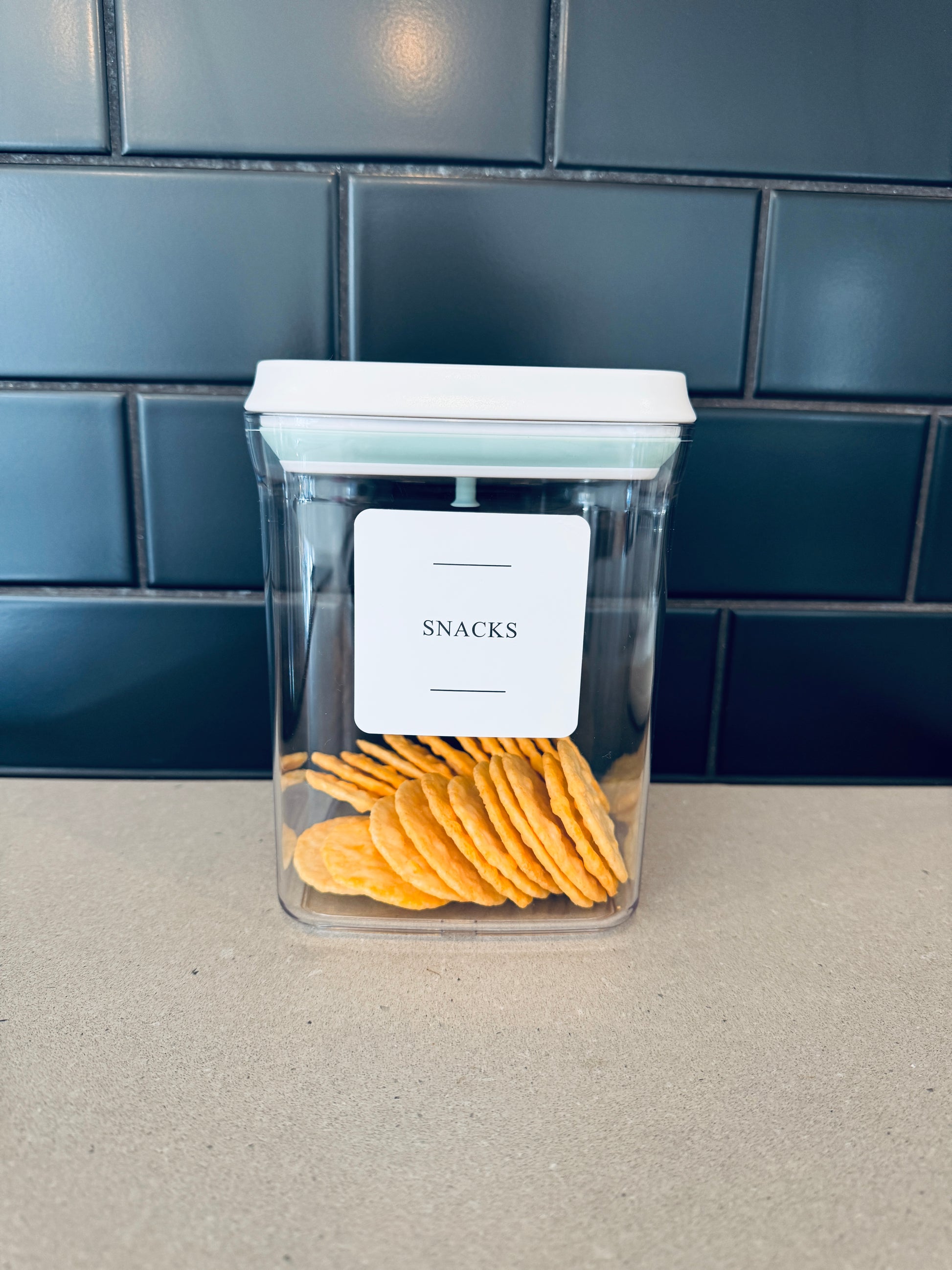 Clear air tight container with crackers labeled 'SNACKS' on a tiled wall background