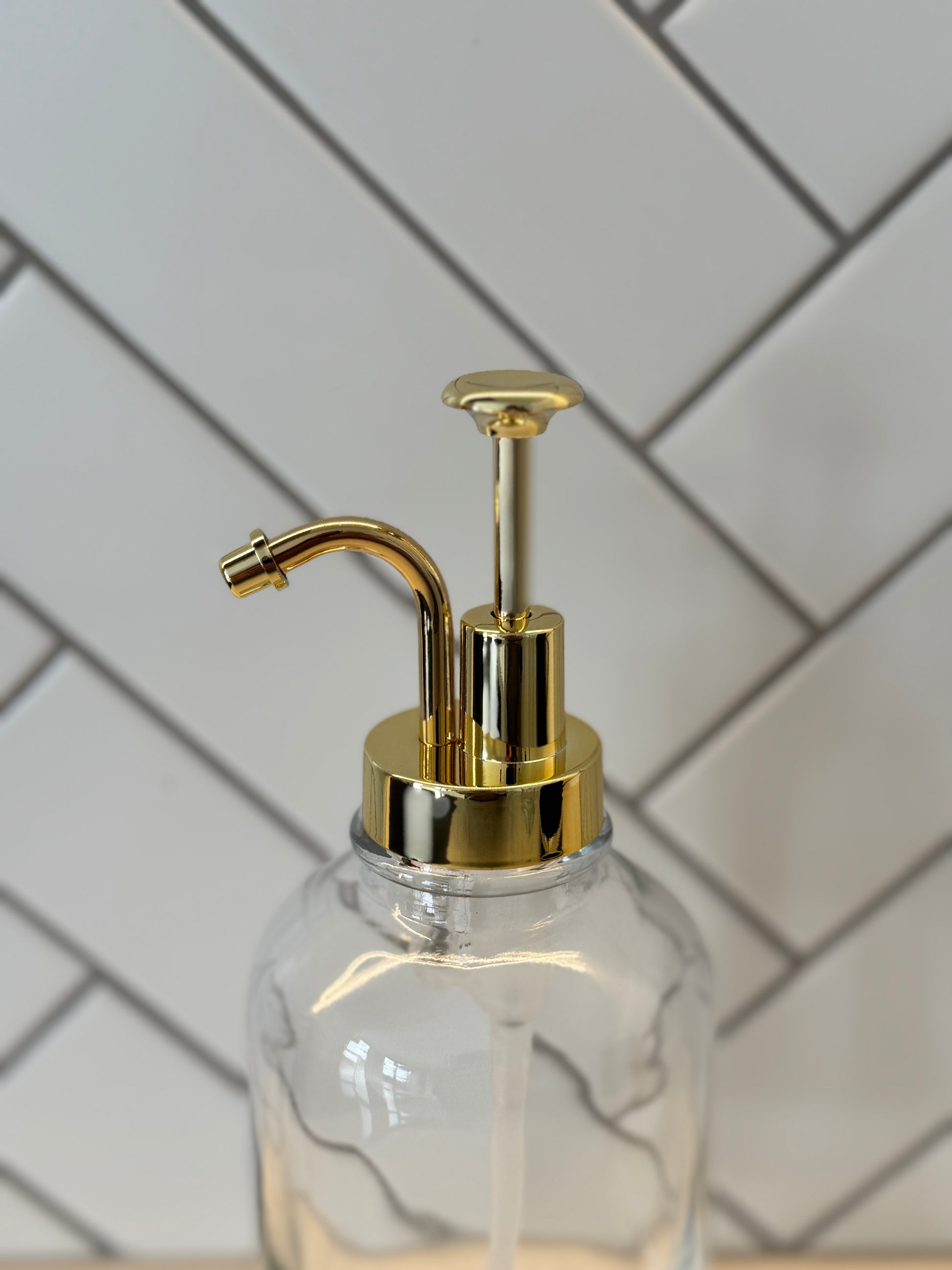 Clear glass syrup dispenser with gold pump against a white tiled wall.