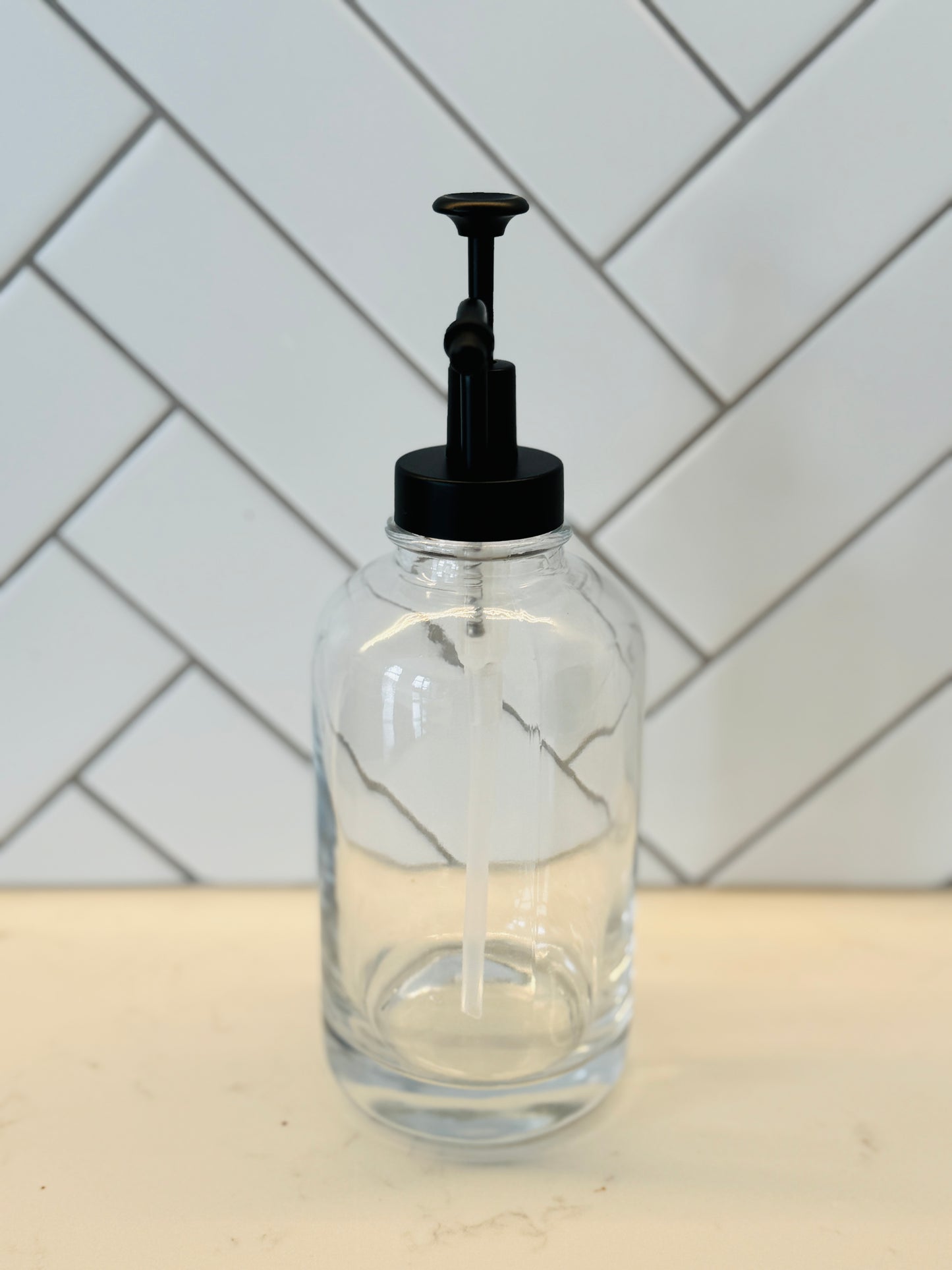 Clear glass bottle with black pump dispenser on a kitchen counter with tiled wall background