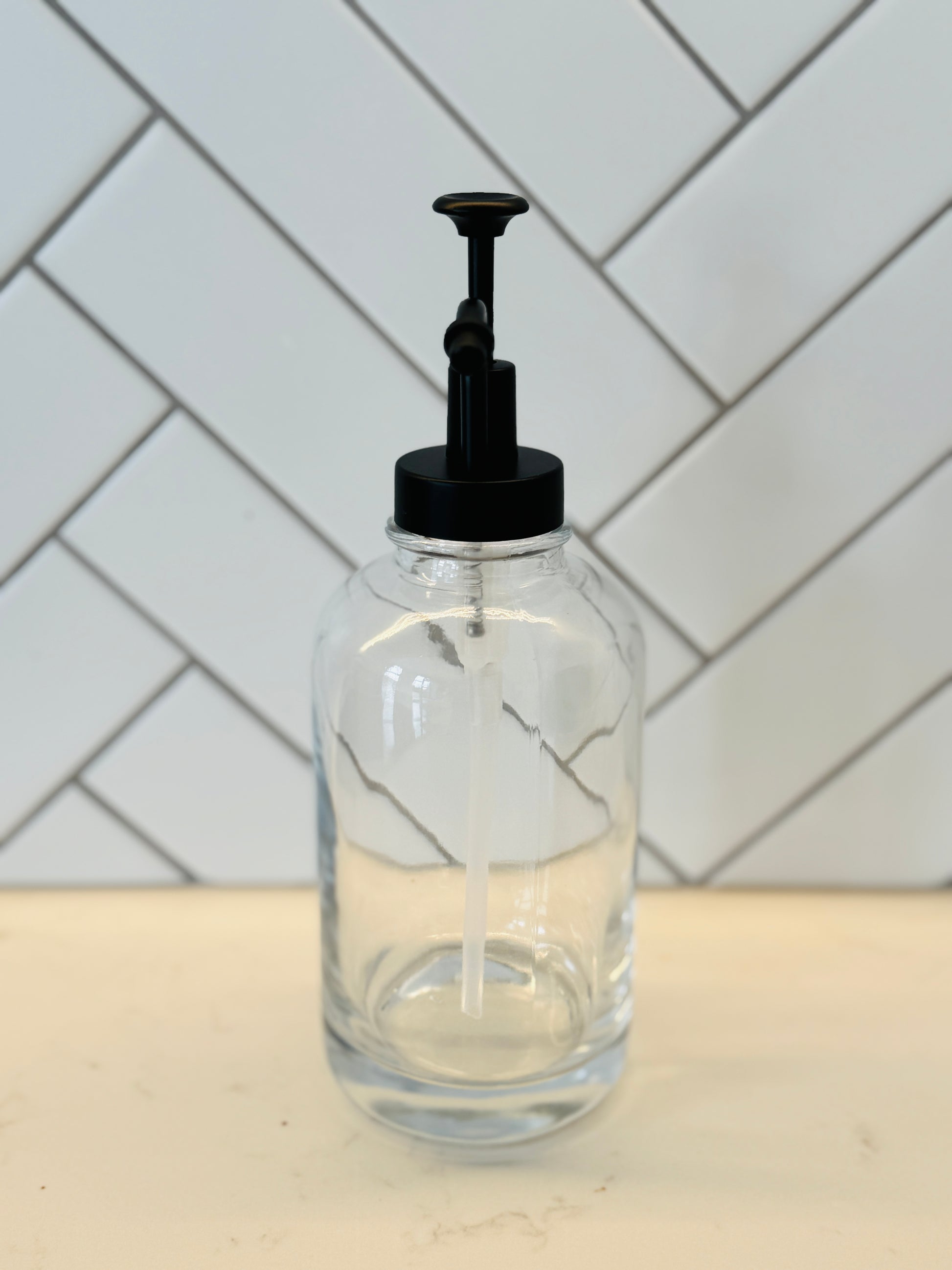 Clear glass bottle with black pump dispenser on a kitchen counter with tiled wall background