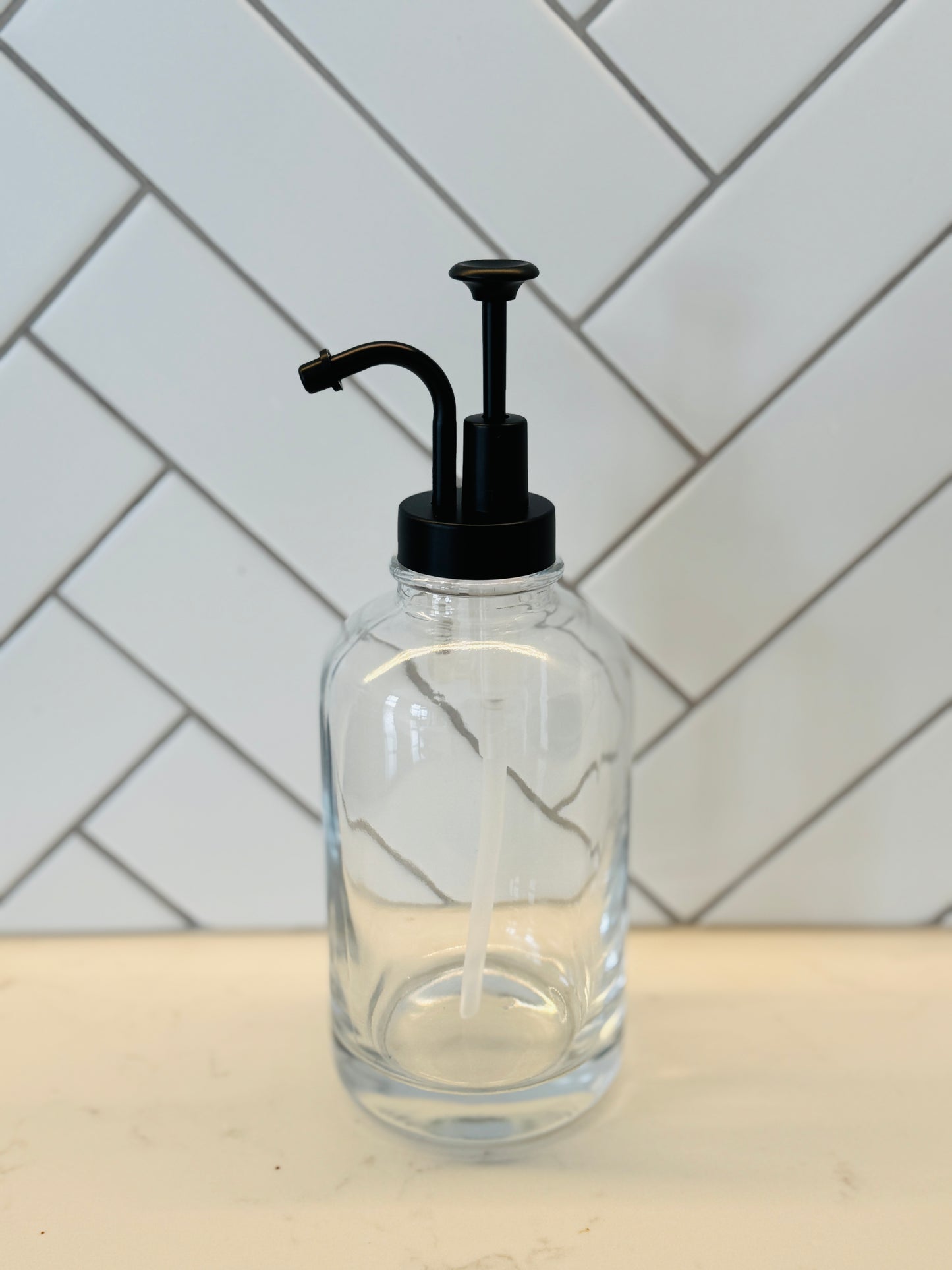 Clear glass bottle with black pump on a white countertop with a tiled wall background