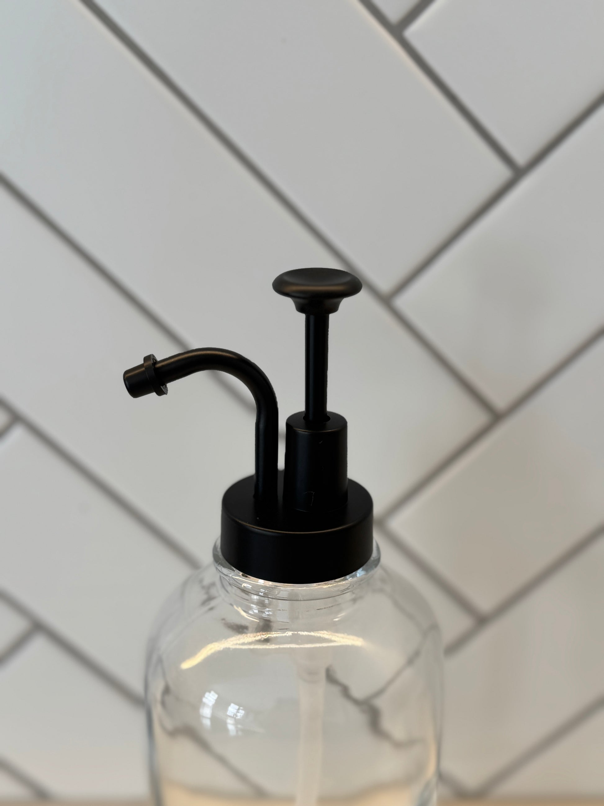 Clear syrup dispenser with black pump against a tiled wall background