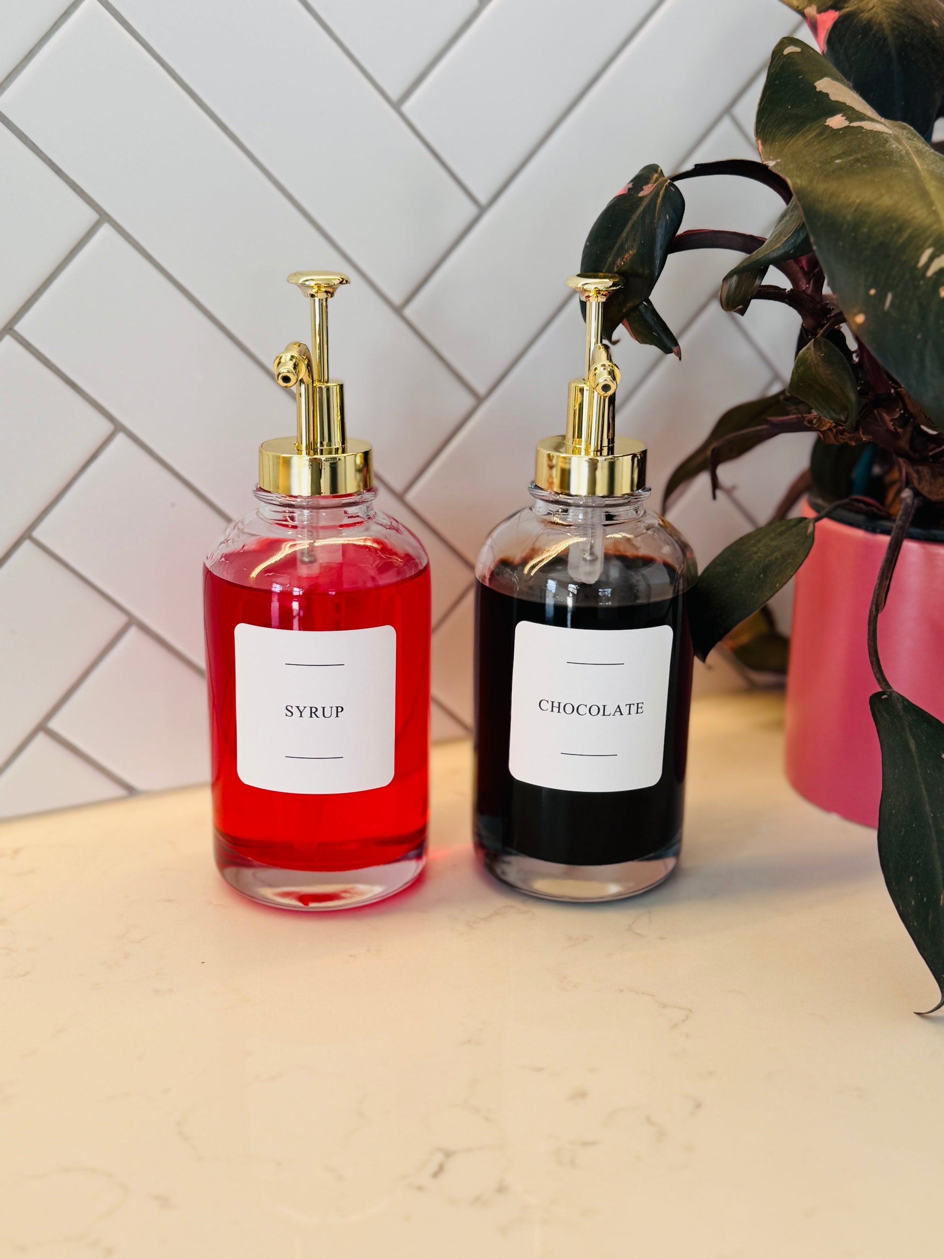 Two glass bottles labeled 'Syrup' and 'Chocolate' on a light surface with a white tiled wall background.