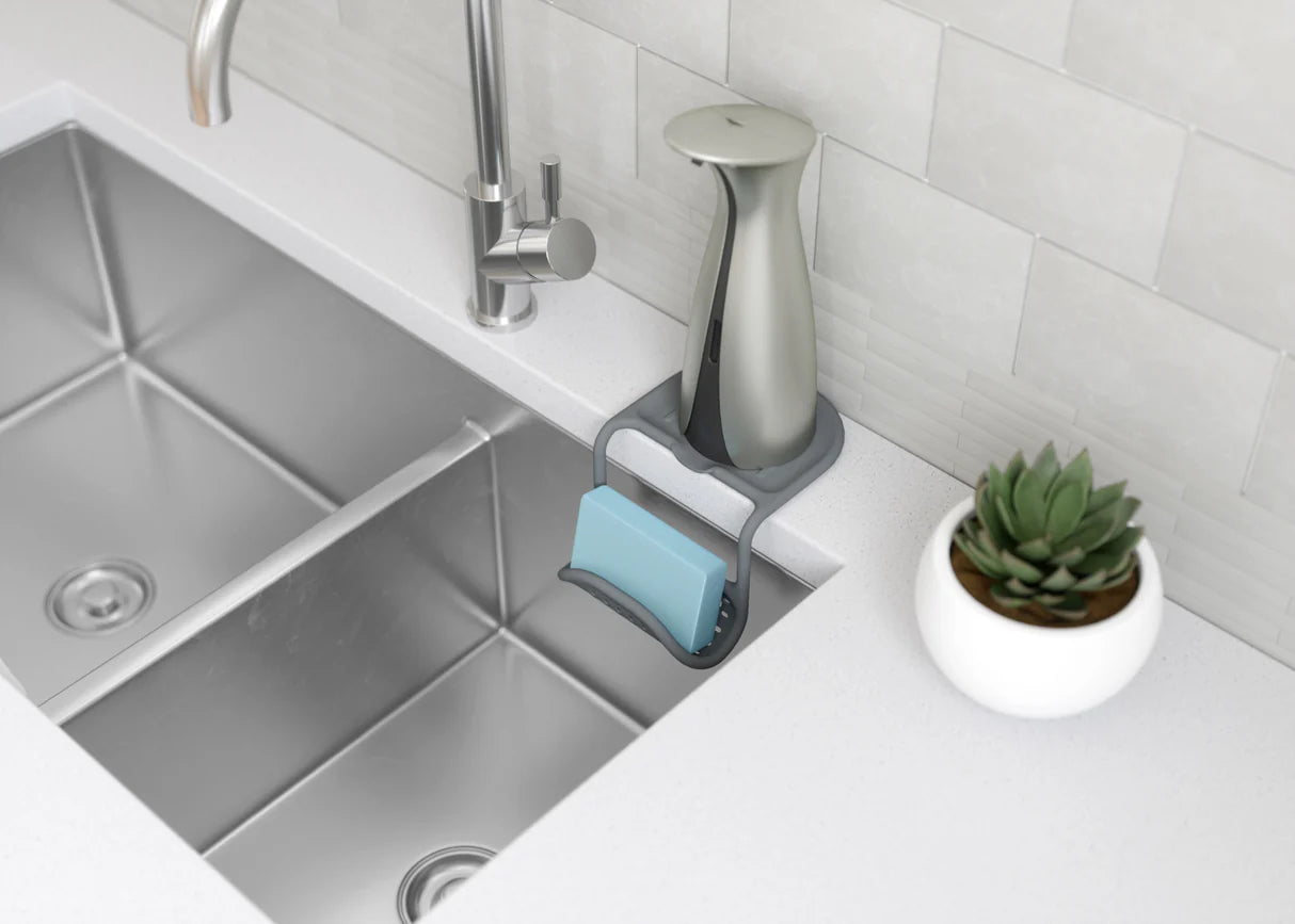 Kitchen sink area with soap dispenser, sponge, and small plant on a white countertop.