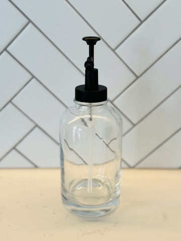 Clear glass bottle with black pump dispenser on a kitchen counter with tiled wall background
